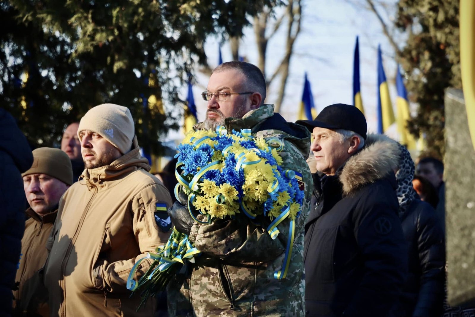 Чоловік у військовій формі тримає букет. На задньому фоні стоять люди на дворі.